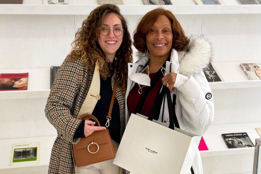 Two smiling women posing together in what appears to a fashion bookshop, one holding a Saint Laurent shopping bag, standing in front of white shelves displaying fashion photography books and magazines.