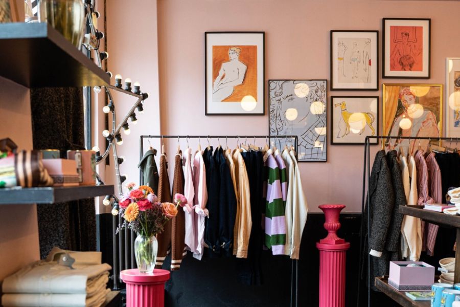Interior of a charming boutique with dusty pink walls, clothing rails displaying jackets and tops, eclectic framed artworks featuring figure illustrations, globe pendant lights, pink pedestal tables, and a vase of fresh flowers.