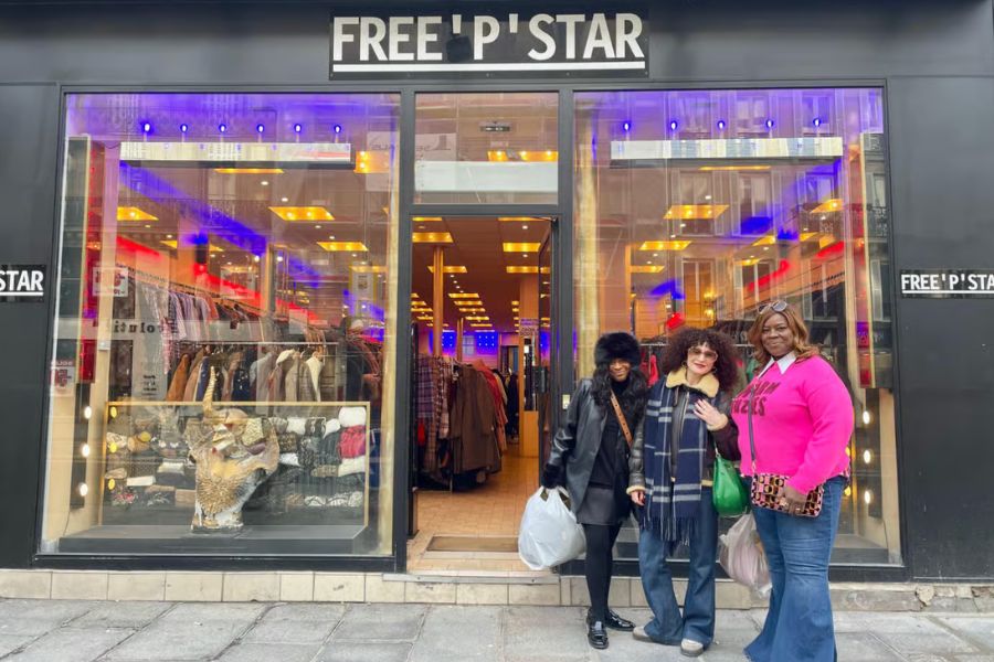 Three women posing and smiling outside Free'P'Star, a well-known Parisian vintage clothing store, with a black shopfront, colourful purple and blue neon lighting visible through the window, and racks of vintage clothes inside.