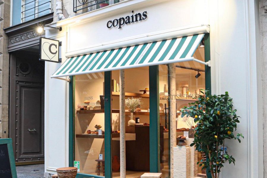 This is an image of the exterior of a cafe where the pain is cream and the accents of the blinds and doorwar are dark green. The cafe's name 'Copains' is in black writing proudly on top.