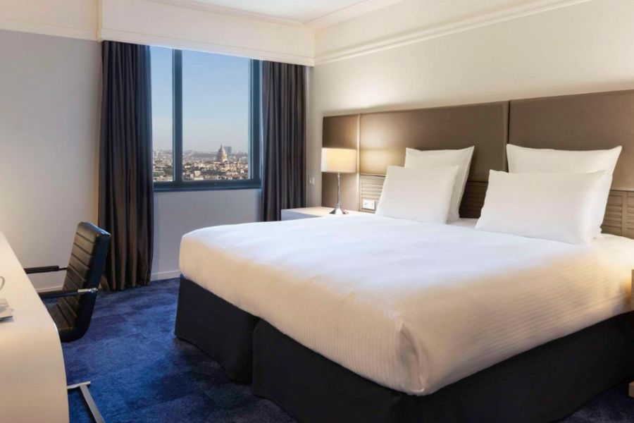 This is an image of a minimalist hotel room with white bedding and a window view of the city.