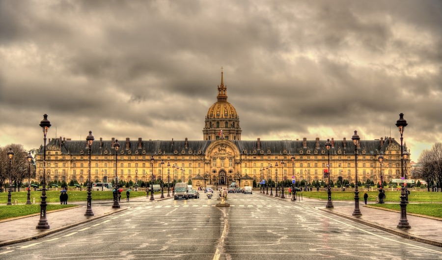This is an image of Les Invalides in Paris. There are lots of clouds in the background but the front of the building looks well lit and warm in tone. It is extremely grand and big.
