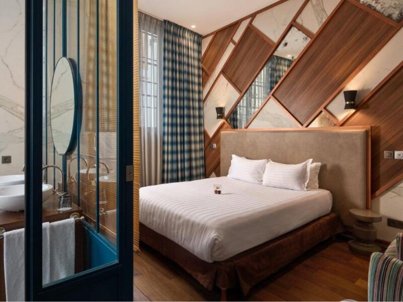 This is an image of a bedroom with an ensuite. There is a massive double bed in the middle and the walls have a cool criss cross pattern of wood, mirror and wallpaper.