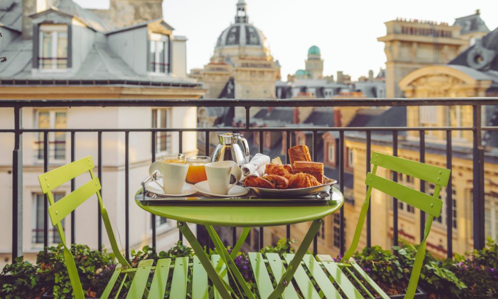 This is an image of a green outdoor table set on a balcony with pastries on it and a nice view of Paris.
