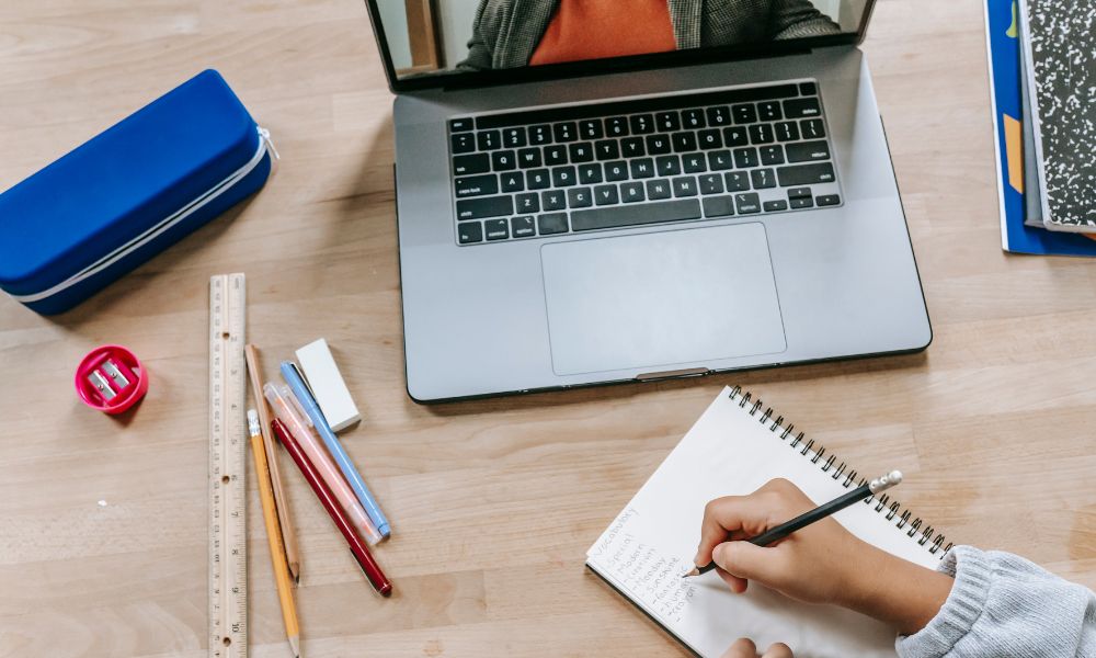 This is an image of a laptop on the table with pens and a pencil case, plus someone writing in a notebook.