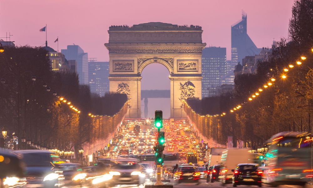This is an image of the Champs Elysees with a view of the Arc De Triomphe at the end of the street. There are cars going left and right and it is very busy.
