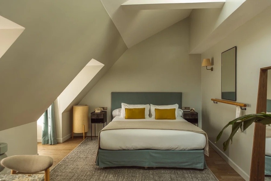 A blue bed, with white line and yellow pillows in a spacious room with hardwood floors.