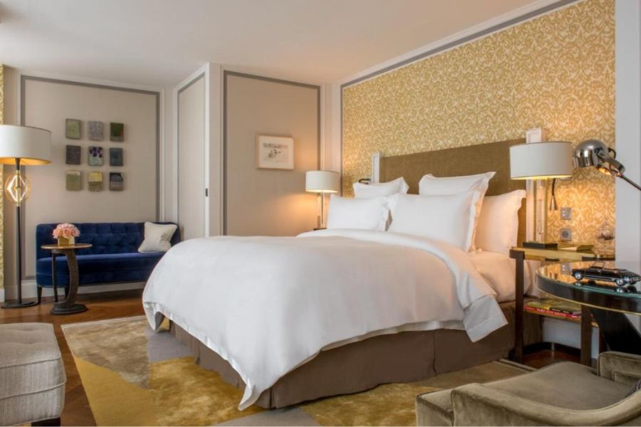 Luxurious hotel room with gold damask feature wall, crisp white bedding, navy blue velvet sofa, gold-toned furnishings, and warm ambient lighting