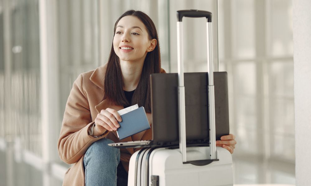 This is an image of a girl sitting down on the floor with her passport and laptop on a small rolling suitcase.