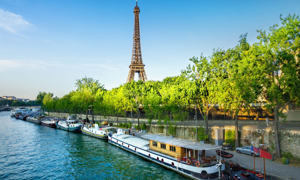 This is an image of the Seine on a beautiful sunny day with the boats moored on the side of the river, the Eiffel Tower in the beckground and big, luscious trees on the riverbank.