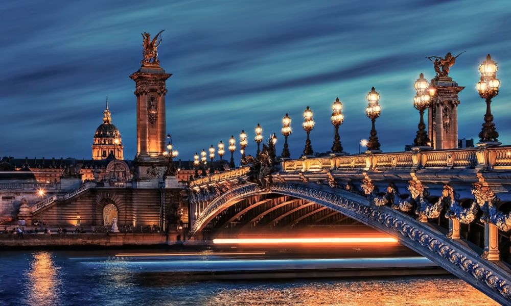 This is an image of a well-lit bridge / area in Paris with the Seine flowing underneath it.