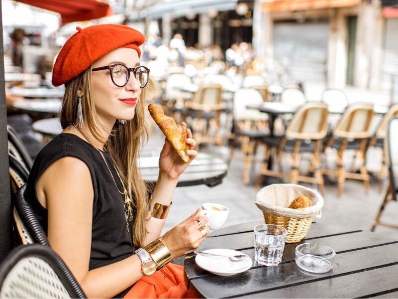 This is an image of a woman in a beret drinking a coffee and eating a croissant.