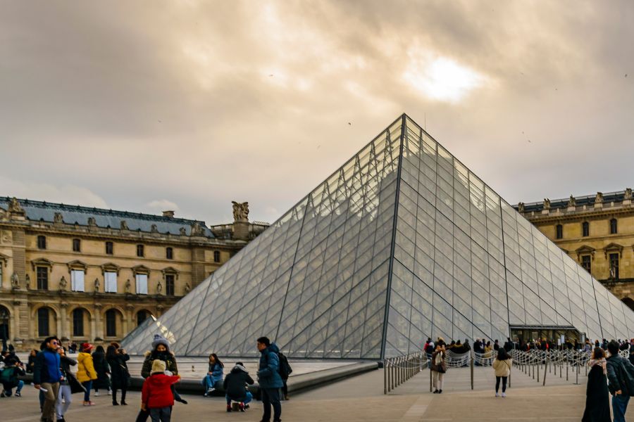 This is an image of the Louvre at sunset with people milling around the area in front taking photos and posing.
