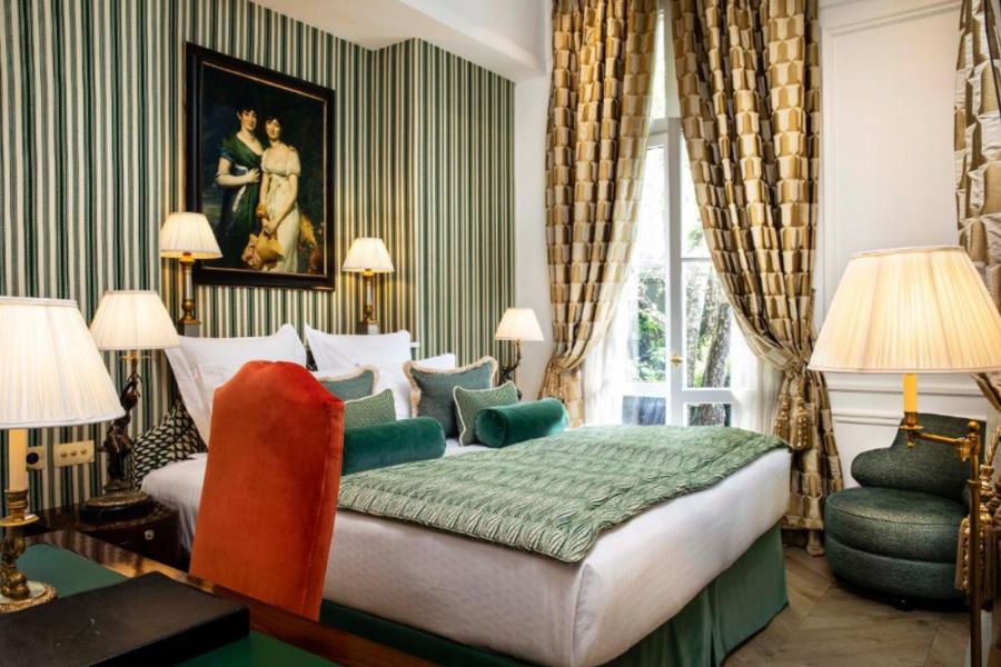 Opulent hotel room with green and white striped wallpaper, a classical oil portrait above the bed, emerald green soft furnishings, and floor-length geometric curtains