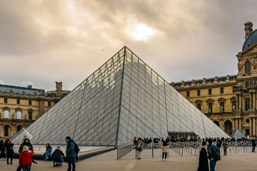 This is an image of The Louvre with people milling around outside and a speck of sun coming through from the clouds.