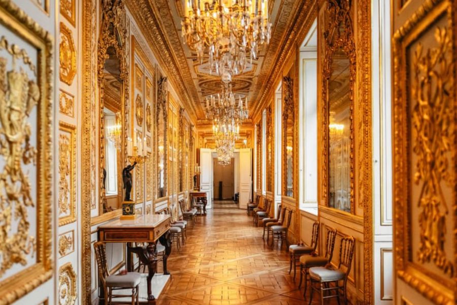 This is an image of the inside of a regal hotel with lots of gold detailing and chandeliers guiding down the hallway.