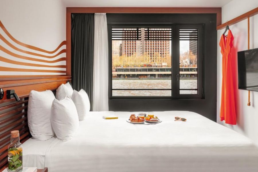 A hotel room with a river view, bold terracotta and black interiors, white bedding and an open hanging rail with an orange dress