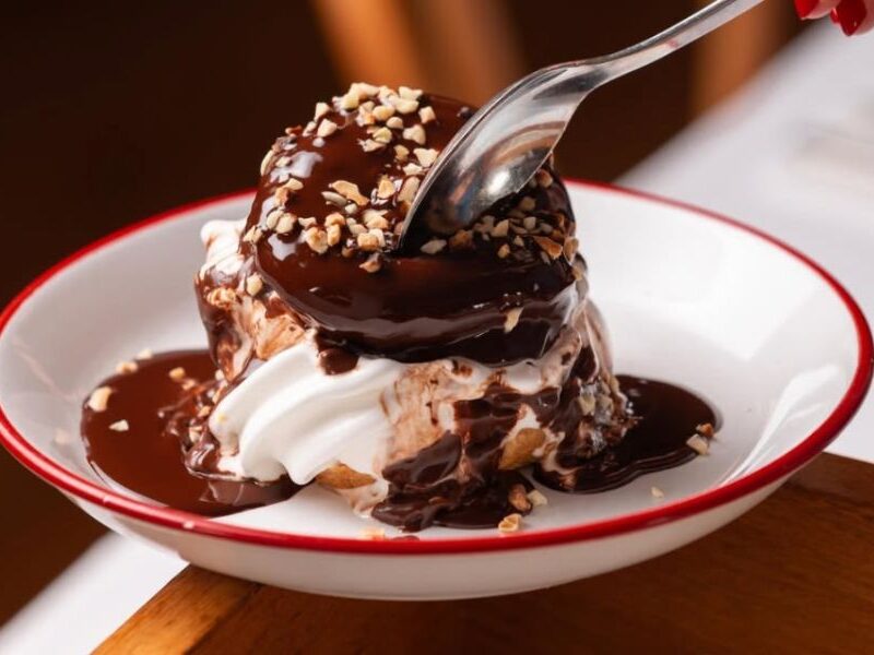 This is an image of a choux pastry with soft serve in the middle and hot chocolate sauce on top, alongside hazelnuts.