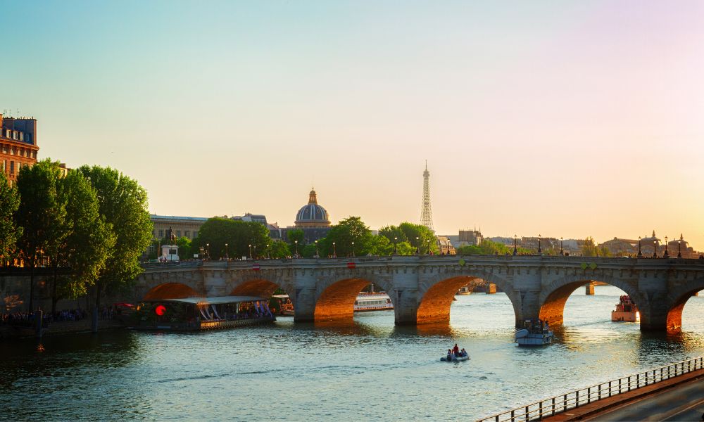 This is an image of a bridge going over the Seine River with the Eiffel Tower in the distance. There is a beautiful golden light reflecting off the water, which shows it's a great place to watch the sunset in Paris.