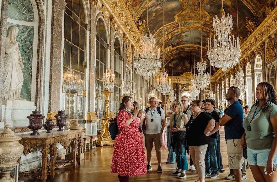 The picture shows the inside of Versailles, with gold details and paintings, along with a tour guide explaining to a group of people.