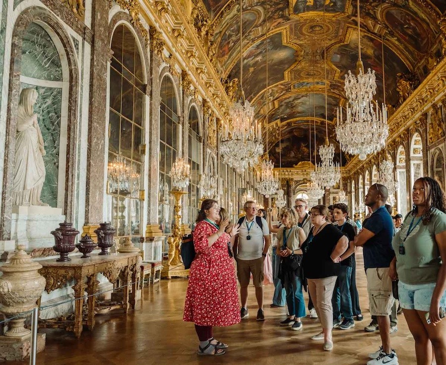 The picture shows the inside of Versailles, with gold details and paintings, along with a tour guide explaining to a group of people.