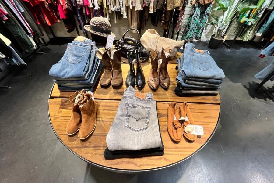 This is an image of a small vintage shop with plenty of clothes neatly hung around and a table with a nice little presentation of shoes and folded jeans.