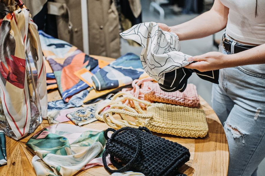 This is an image of a lady touching a scarf in a shop. Surrounding her, is a bunch of other items.