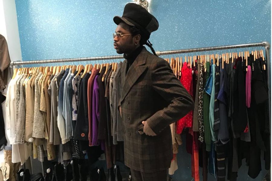 This is an image of a man standing in front of racks of vintage clothing trying on a suit.
