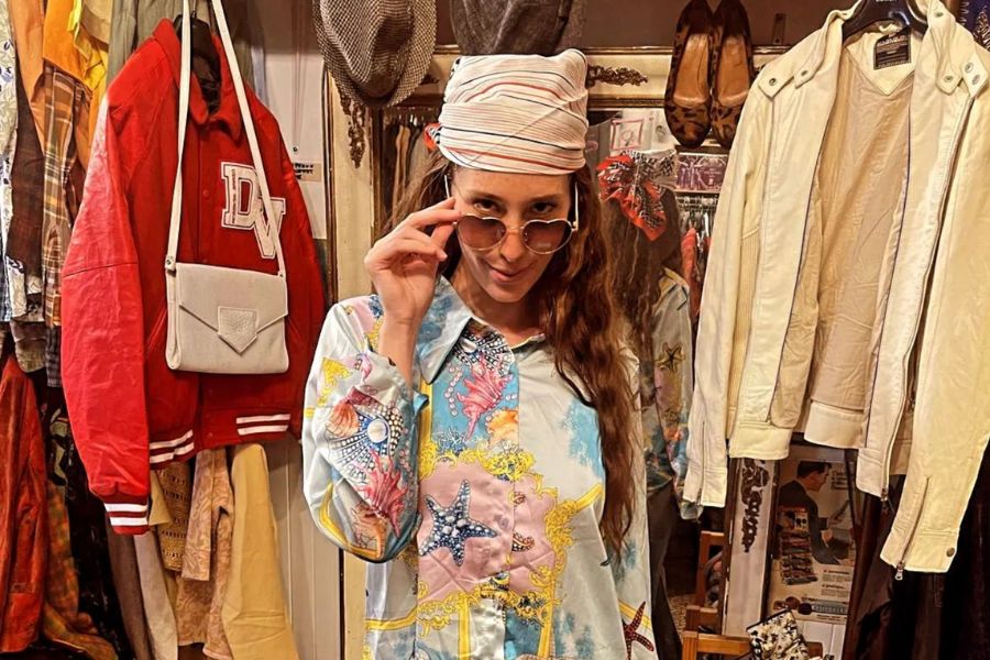 This is an image of a lady wearing cool clothes and tipping her sunglasses at the camera surrounded by clothes.