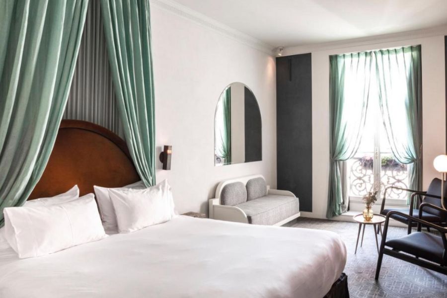 This is an image of a big, minimalist room in a Paris hotel with white sheets on all the bedding and decor, with a single accent drape over the bed that is green.