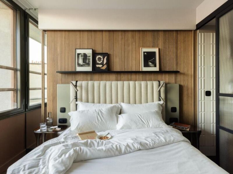 This is an image of a minimalist hotel room that looks more like a nice bedroom than accommodation. The sheets are white and there is a wooden feature wall.