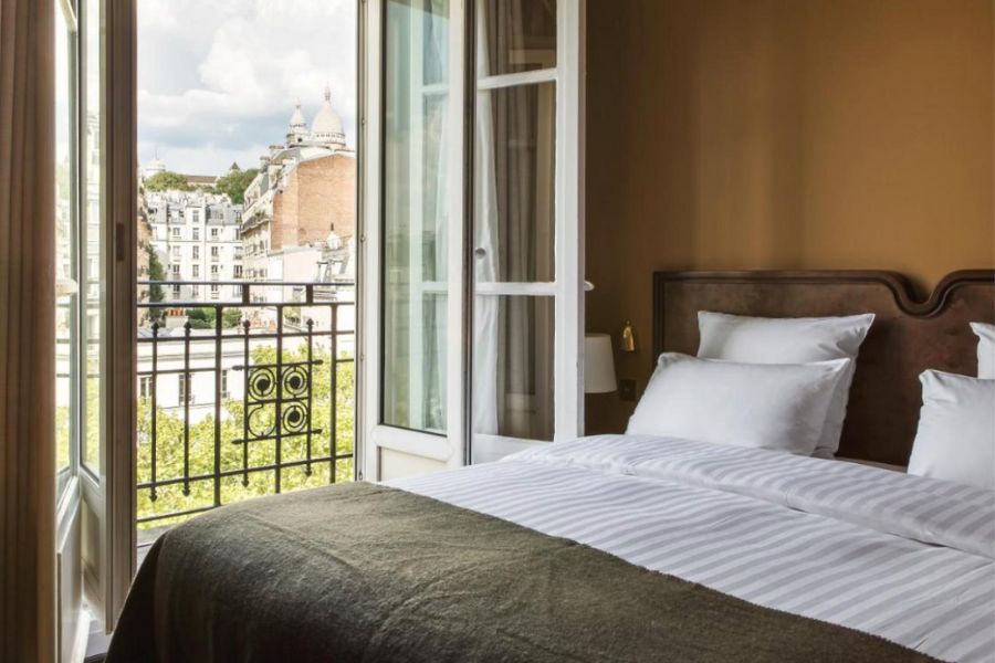 This is an image of a nice minimalist hotel room with a double bed, burnt orange walls and a wonderful balcony overlooking a view of Paris.