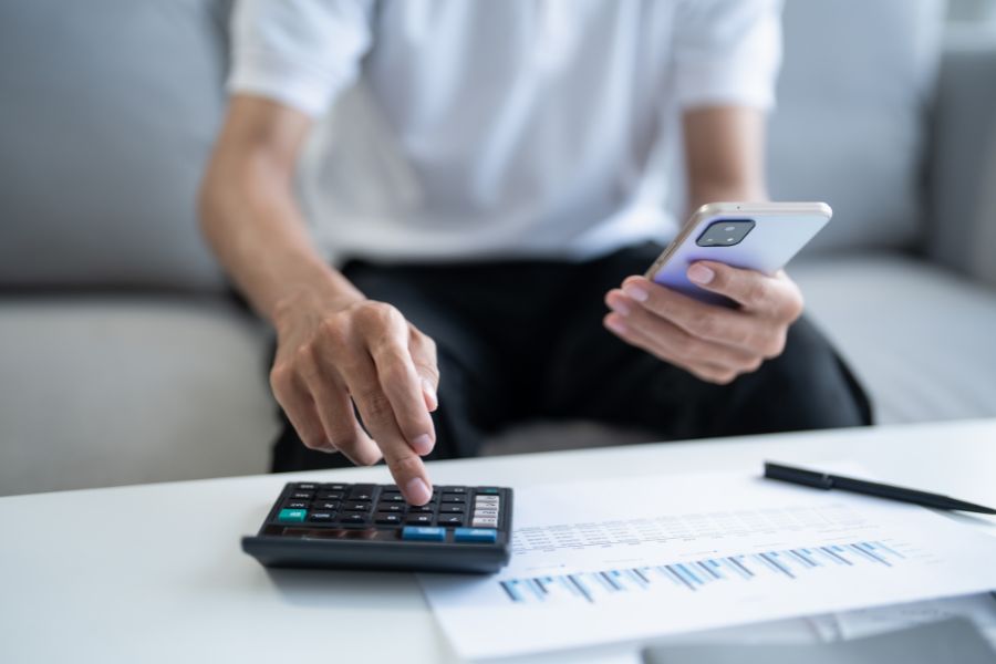 A person sitting on a sofa uses a calculator with one hand while holding a smartphone in the other, with printed financial documents on the table in front of them.