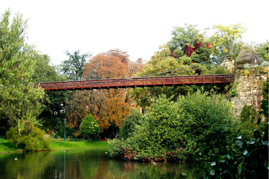 This is a picture of a big green space with a lake and a bridge on top in Paris.