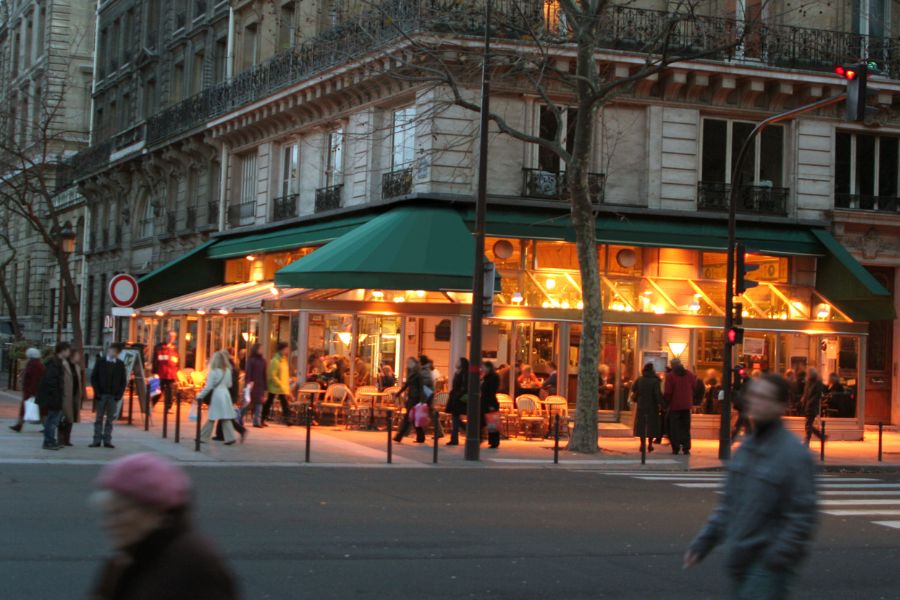 This is a picture of the lively outdoor cafe in the early evening. The lights are on and people are walking around outside.