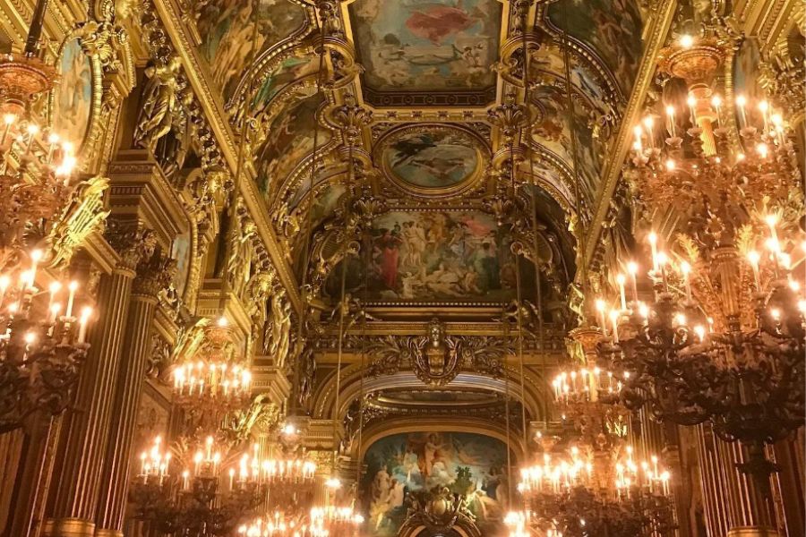 This is an image of the gold interior of the Opera Garnier. There are paintings on the roof and chandeliers around.