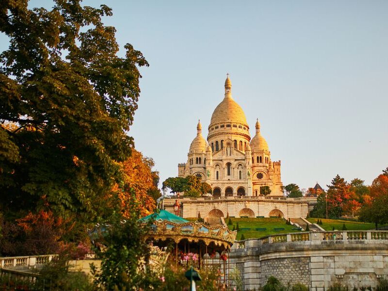 This is an image of the Sacre-Coeur on a hill during sunset time with greenery surrounding.