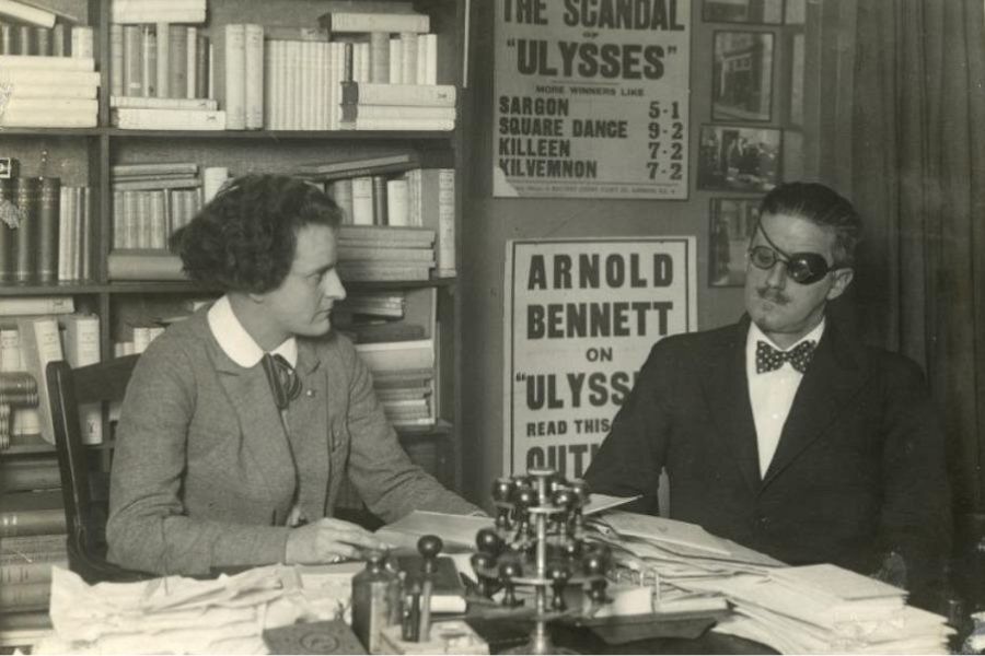 Best literary spots in Paris: This is an image of two people from literary history sitting and conversing with one another at a table surrounded by books and old posters. The picture is black and white.
