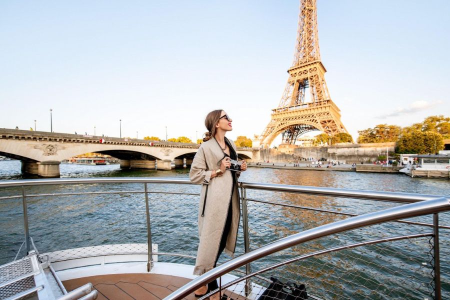 This is an image of a boat on the Seine with a lady looking out to the distance where the Eiffel Tower sits. She is holding a camera.