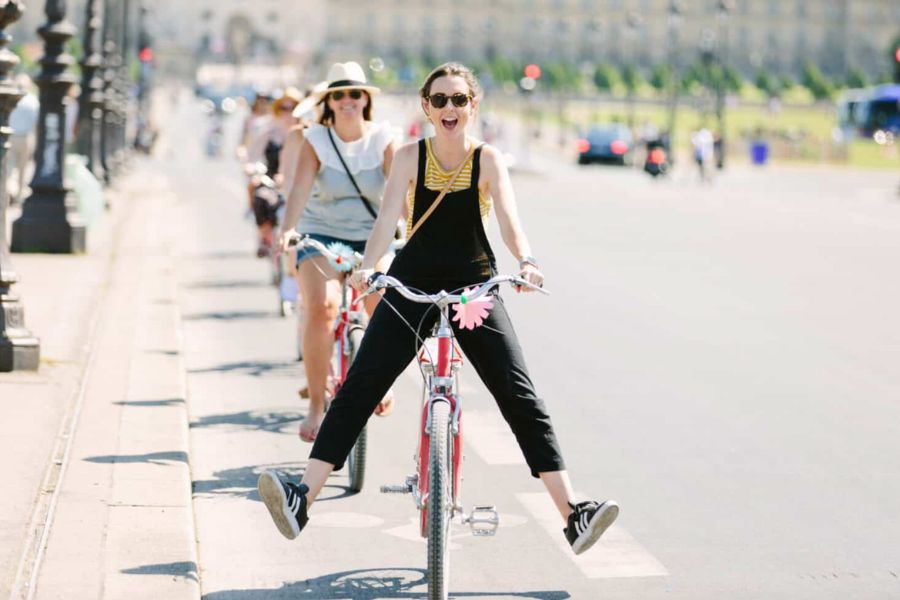 This is a tour group of people riding down the street on bikes. The one at the front has an excited expression and she is dangling her legs away from the pedals.