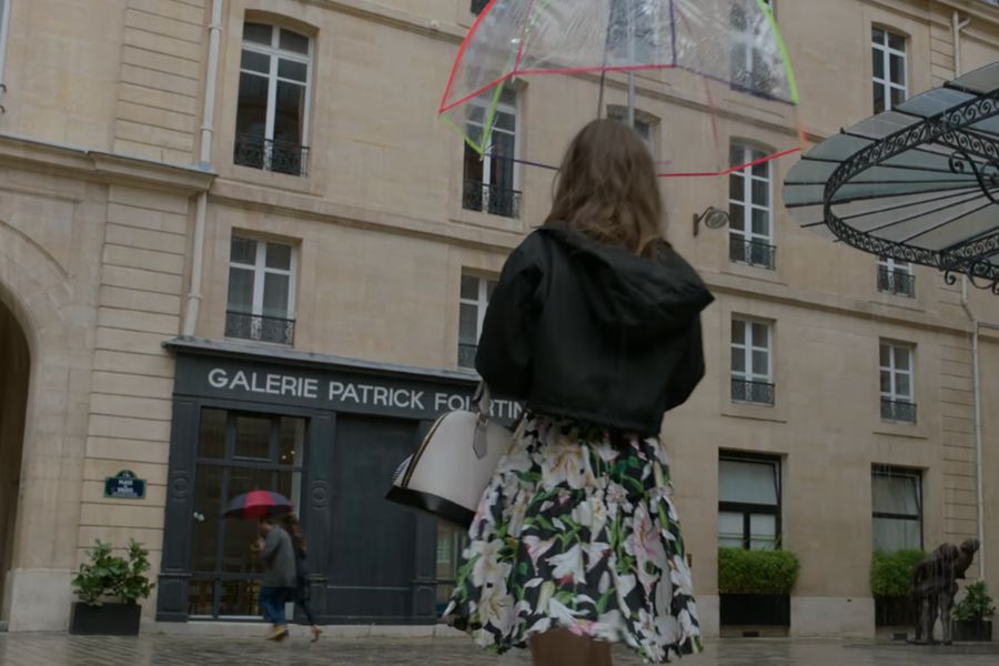 Emily in Paris filming locations: This is an image of Emily walking to the front of her office in the rain. She is holding an umbrella.