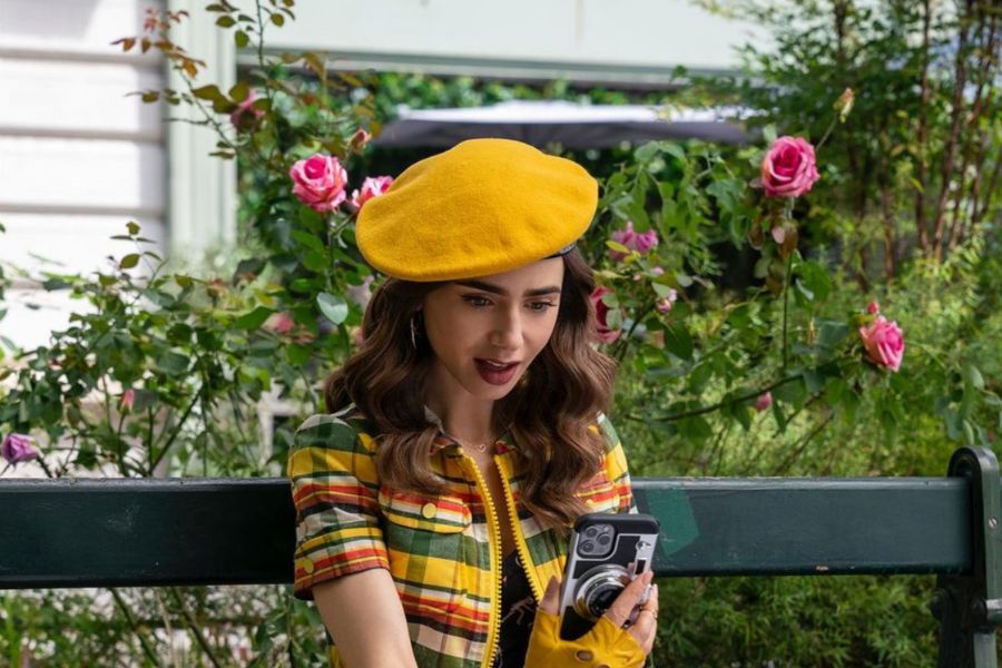 This is an image of Emily sitting on a park bench in front of a rose bush. She seems to be on FaceTime on her phone.