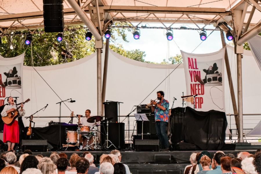 Musicians performing on an outdoor stage in front of a seated crowd, with an "All You Need Is FIP" banner