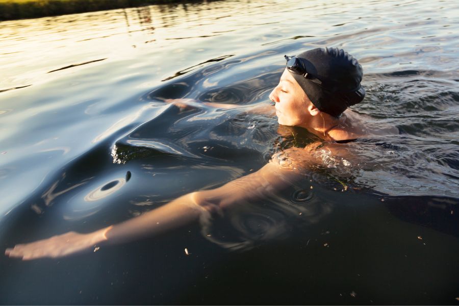 A swimmer in a black cap and goggles swimming in open water, lit by golden evening light