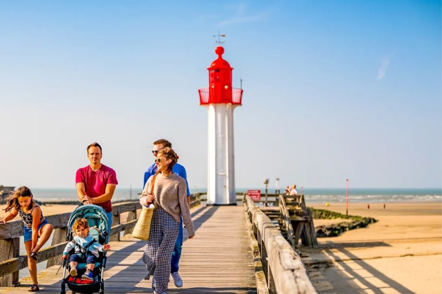 A family walking along a wooden pier towards a red and white lighthouse on the beach