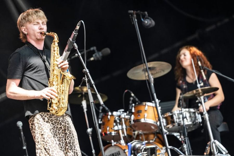 A saxophonist performs on stage with a drummer behind him at an outdoor festival