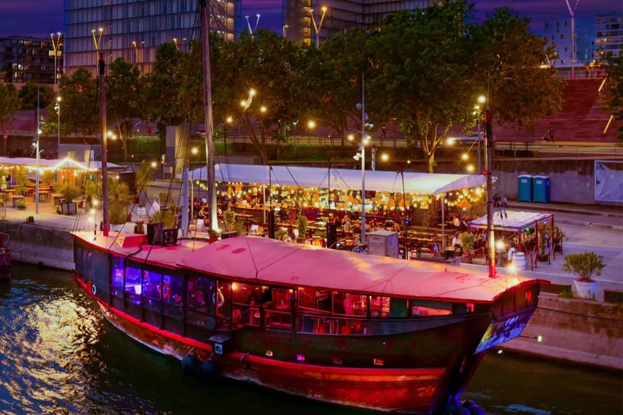 A red-lit boat moored on the Seine at night, with a lively riverside bar lit by string lights