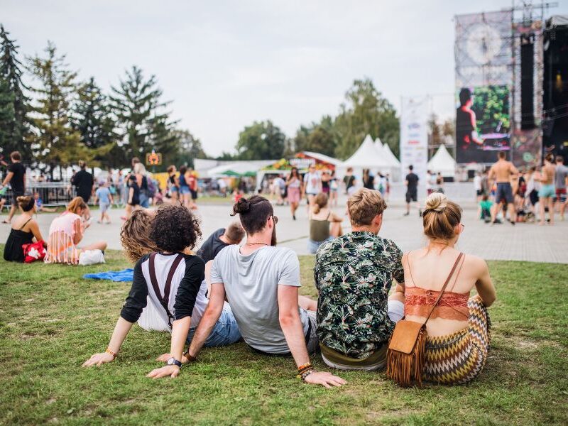 This is an image of a summer festival where four friends are sitting down watching the stage and all the stalls around it.