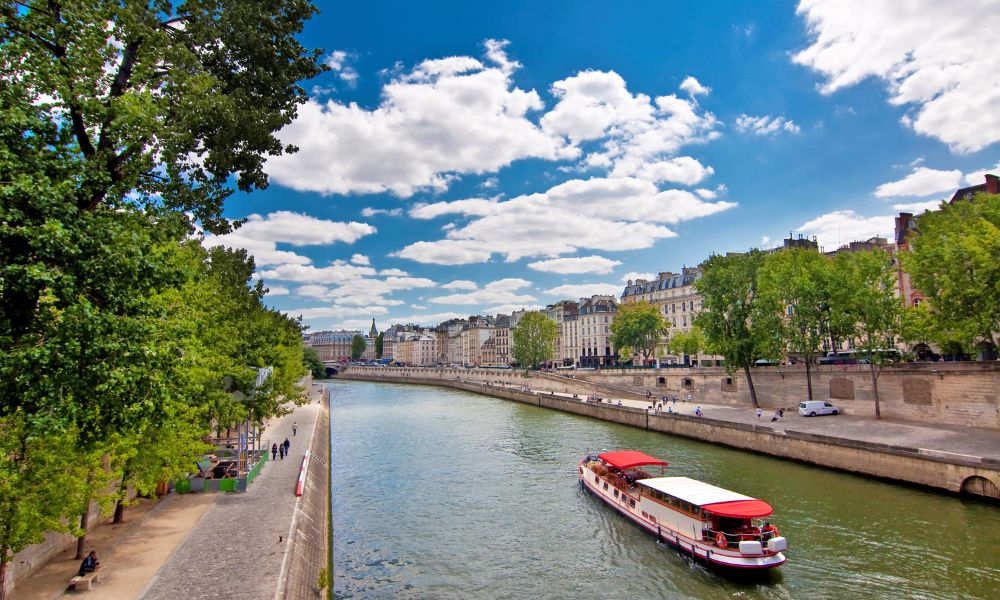 This is an image of a boat riding through the Seine River with blue skies, lined with green trees.