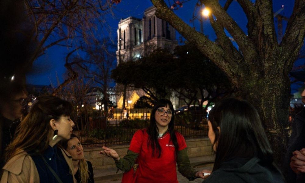 This is an image of a tour in Paris that is running in the evening with a tour guide and group. they are standing in front of a well-lit street in front of the Notre Dame.